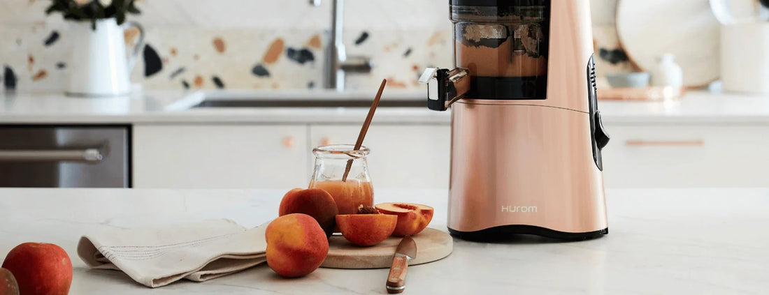 Rose gold Hurom slow juicer on a kitchen counter with peaches and a glass of juice