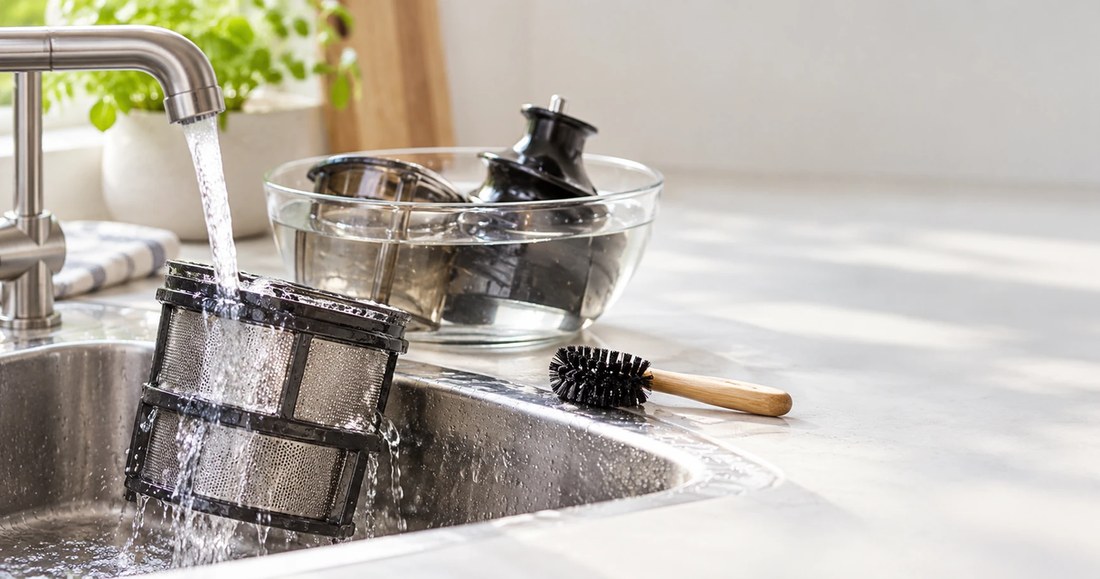 A slow juicer mesh screen under a running tap with a small cleaning brush