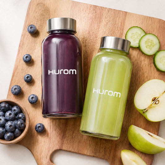 Hurom juice bottles with purple and green drinks on a wooden board with fresh fruits