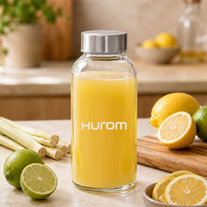 Hurom glass bottle filled with citrus juice surrounded by lemons, limes, and lemongrass